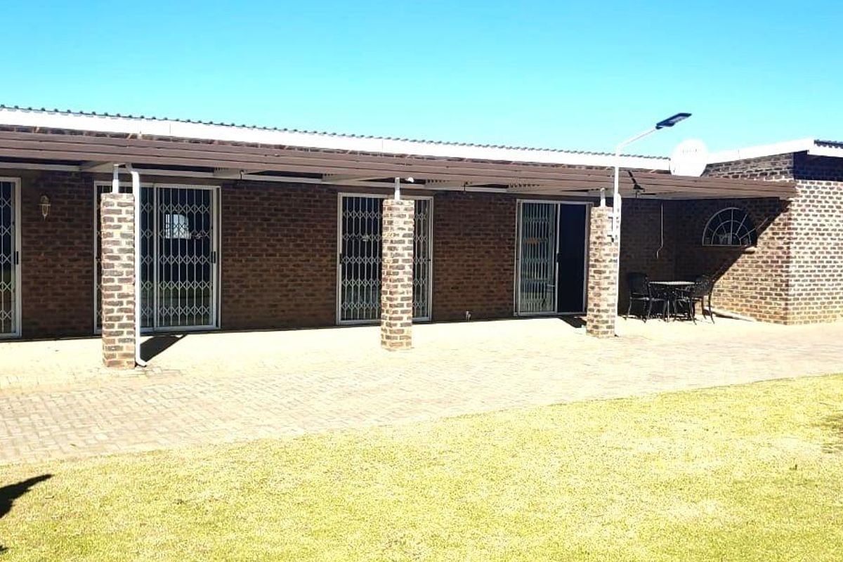 Rear view of the house featuring a spacious paved patio area, covered veranda with brick pillars, and multiple sliding door access points with security gates.