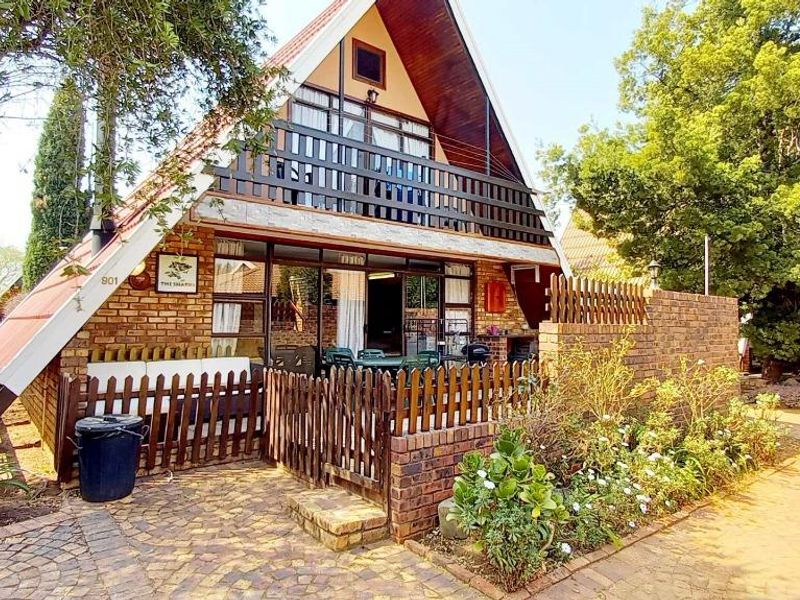 A charming A-frame house featuring a brick facade, a private timber balcony, and a paved entrance area surrounded by established garden landscaping.