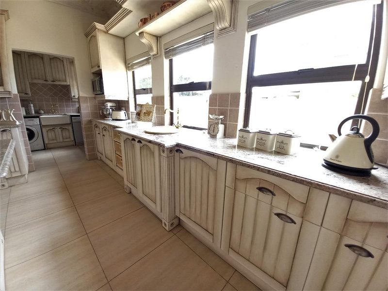 A traditional kitchen featuring cream-toned cabinetry, granite countertops, a farmhouse sink, and tiled flooring, with an adjacent scullery area visible.