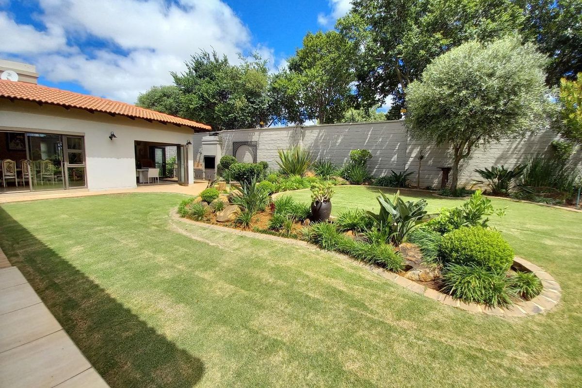 A well-maintained rear garden featuring a manicured lawn, landscaped garden beds, a tiled patio area, and a built-in outdoor braai.