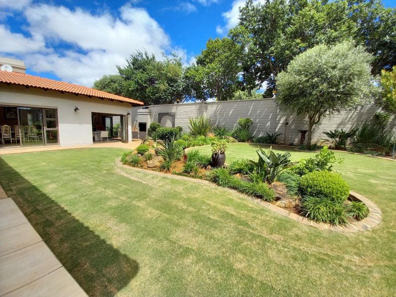 A well-maintained rear garden featuring a manicured lawn, landscaped garden beds, a tiled patio area, and a built-in outdoor braai.