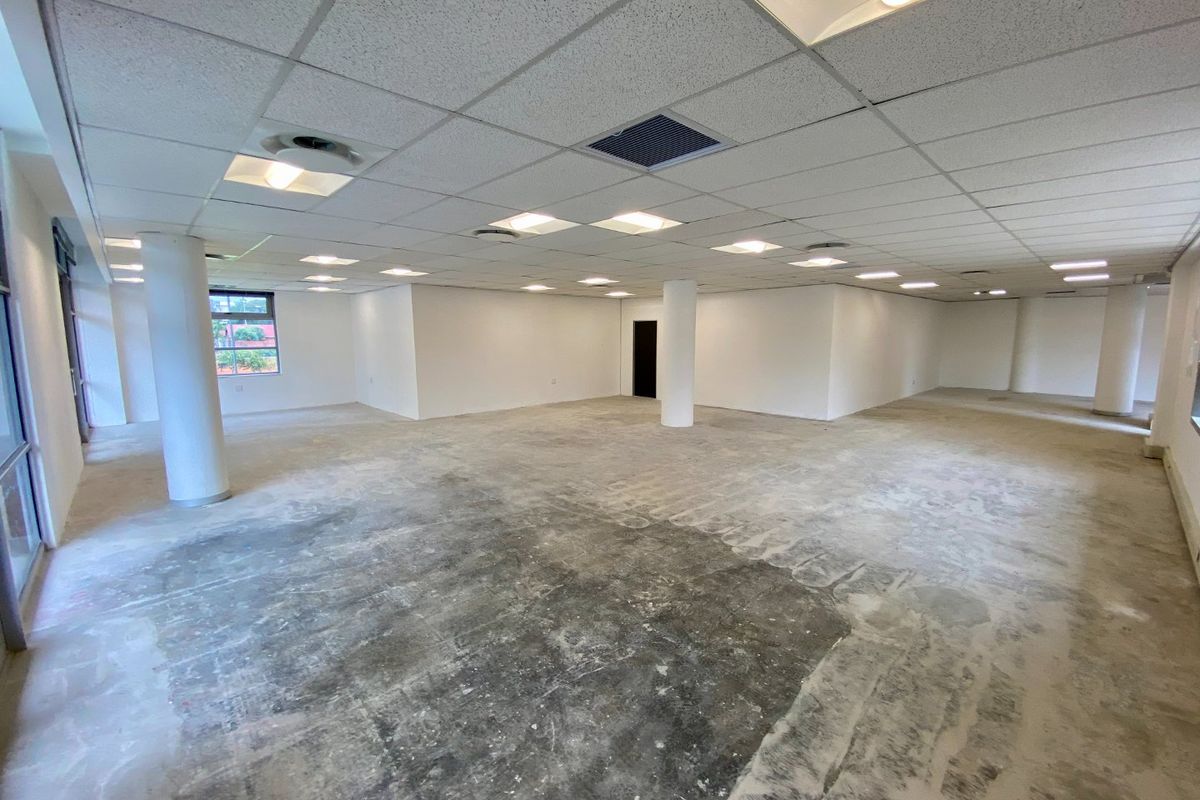 A spacious, open-plan commercial office area featuring a suspended ceiling, recessed lighting, and structural columns, currently awaiting final flooring installation.