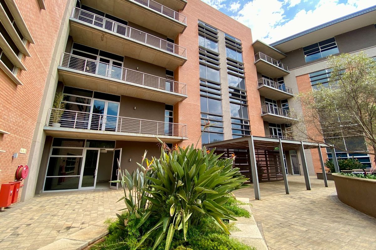 A modern commercial building exterior featuring a paved courtyard, multi-level balconies with metal railings, and large glass windows.