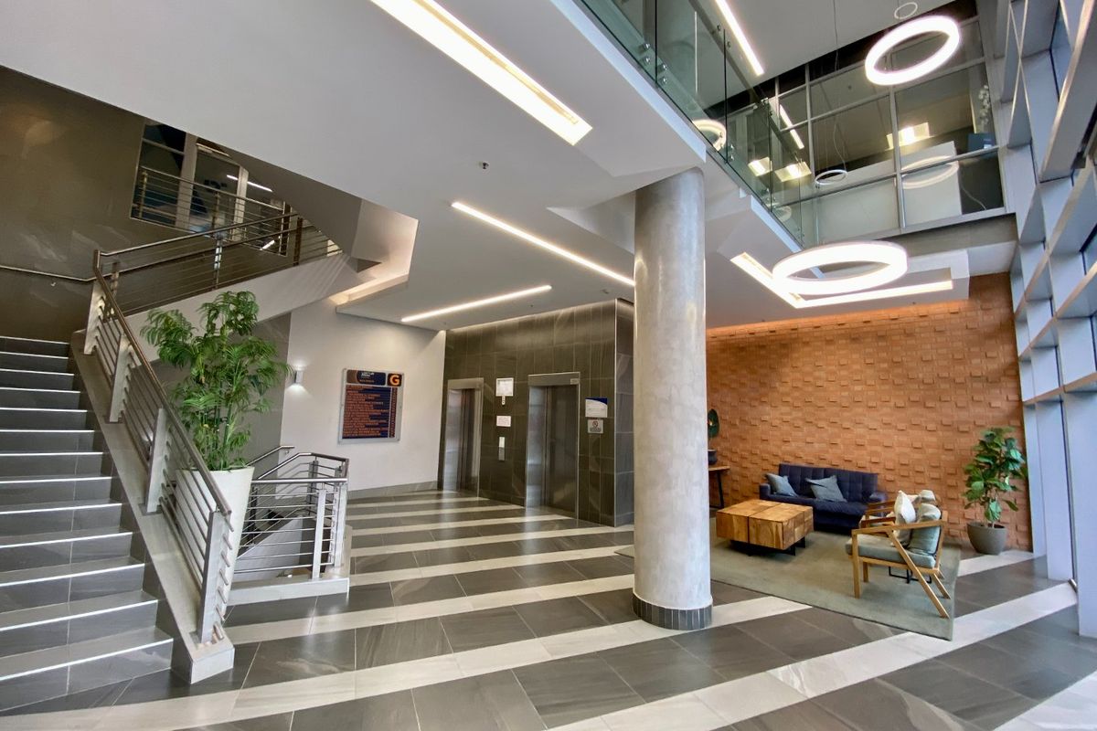 Modern commercial building lobby featuring a grand staircase, elevator access, high ceilings with contemporary lighting, and a textured feature wall.