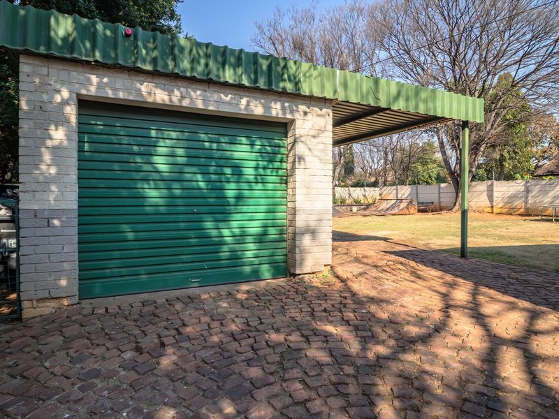A detached garage featuring a green roller shutter door, a covered carport extension, and a brick-paved driveway set within a spacious garden.