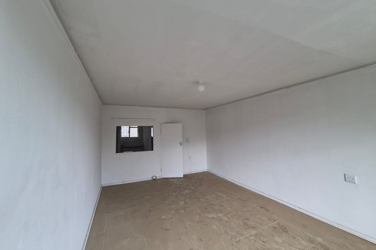 An empty, neutrally painted living area featuring tiled flooring and an interior service window looking into the kitchen space.
