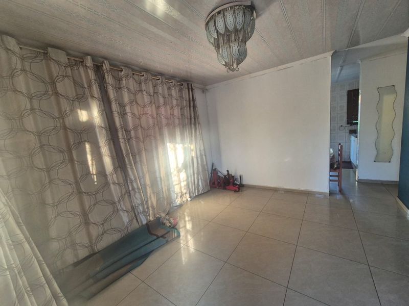 An empty living room featuring tiled flooring and a central ceiling light fixture, with direct access to an adjacent kitchen area.