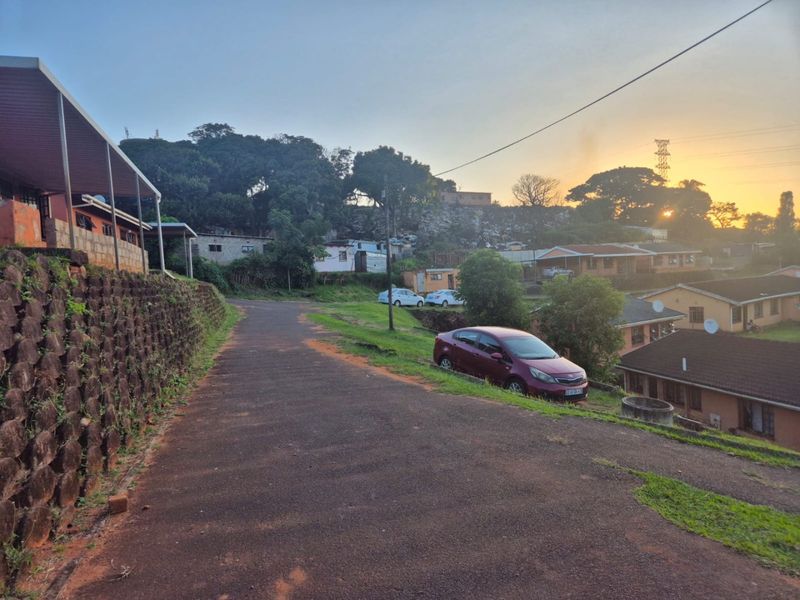 A view of the property approach featuring an asphalt driveway, a stone retaining wall, and surrounding residential buildings at sunset.