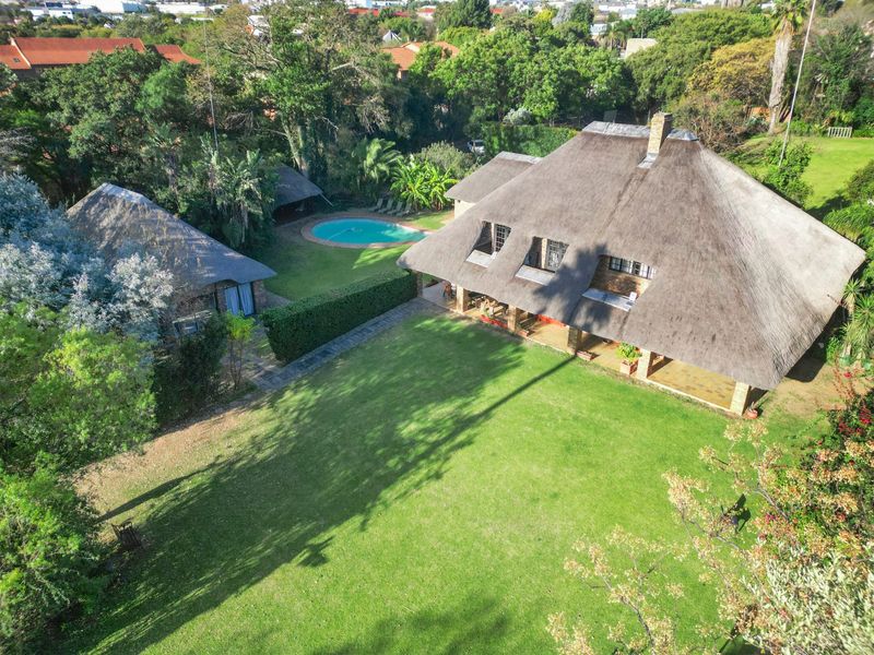Aerial view of a commercial property featuring a large thatched-roof building, expansive lawn, swimming pool, and mature garden surroundings.