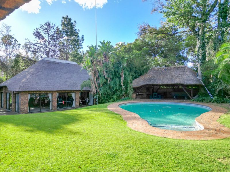 Outdoor entertainment area featuring a swimming pool, landscaped garden, and a thatched-roof building with large sliding glass doors.