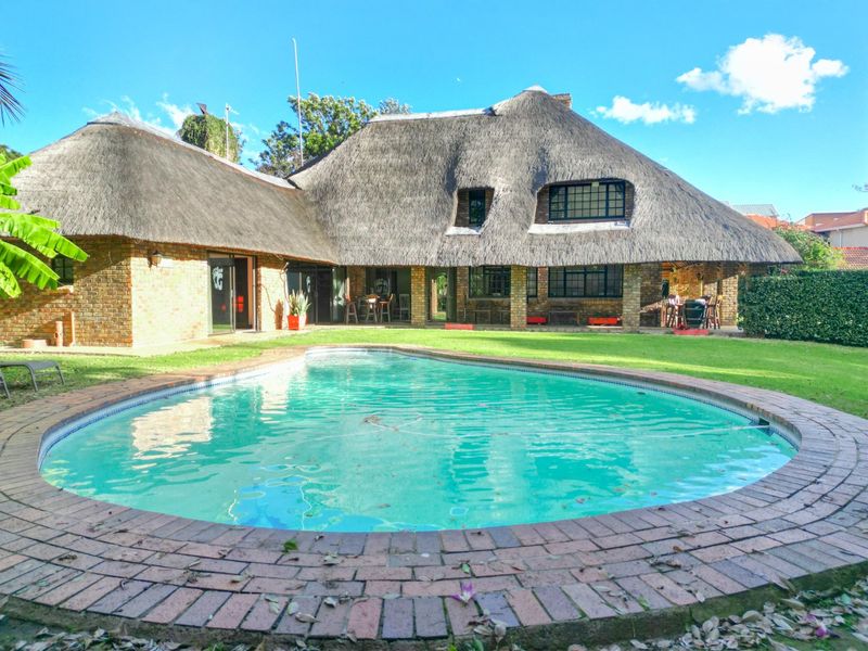 Rear view of a commercial property featuring a large swimming pool with brick coping and a traditional thatched roof building with a covered patio area.