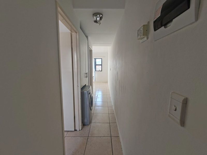 A narrow hallway featuring tiled flooring, built-in cabinetry, and an electrical distribution board, leading towards a windowed area.