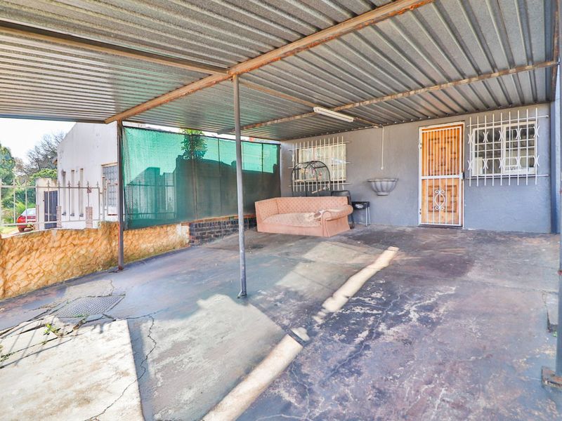 A covered outdoor patio or carport area featuring a corrugated metal roof, concrete flooring, and security bars on windows and doors.