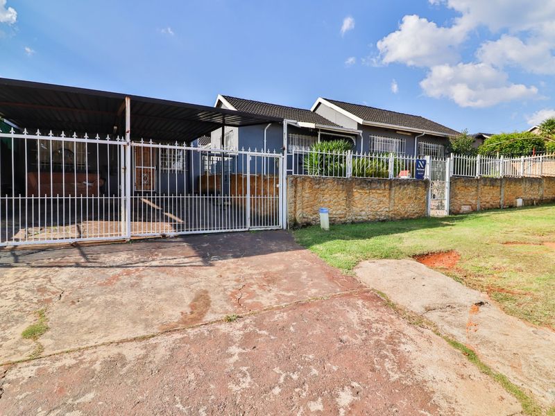 Front exterior view of the property featuring a paved driveway, covered carport, and stone boundary wall with white metal security fencing.