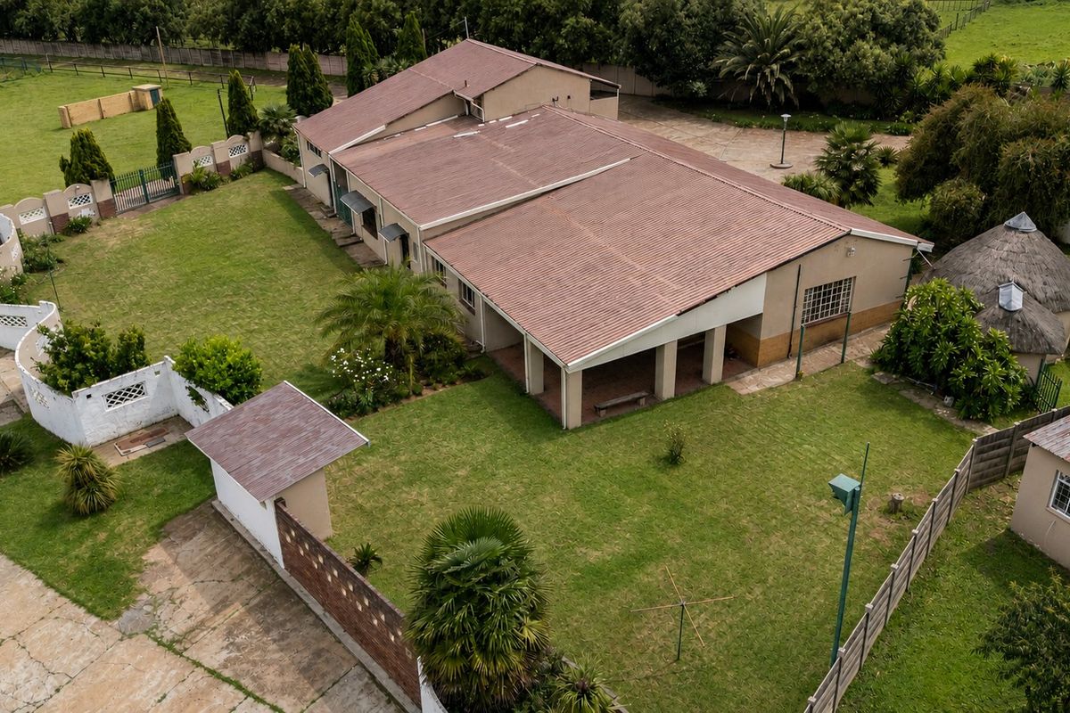 Aerial view of a farm property featuring a main dwelling with a covered patio, expansive lawn, perimeter fencing, and a secondary thatched structure.