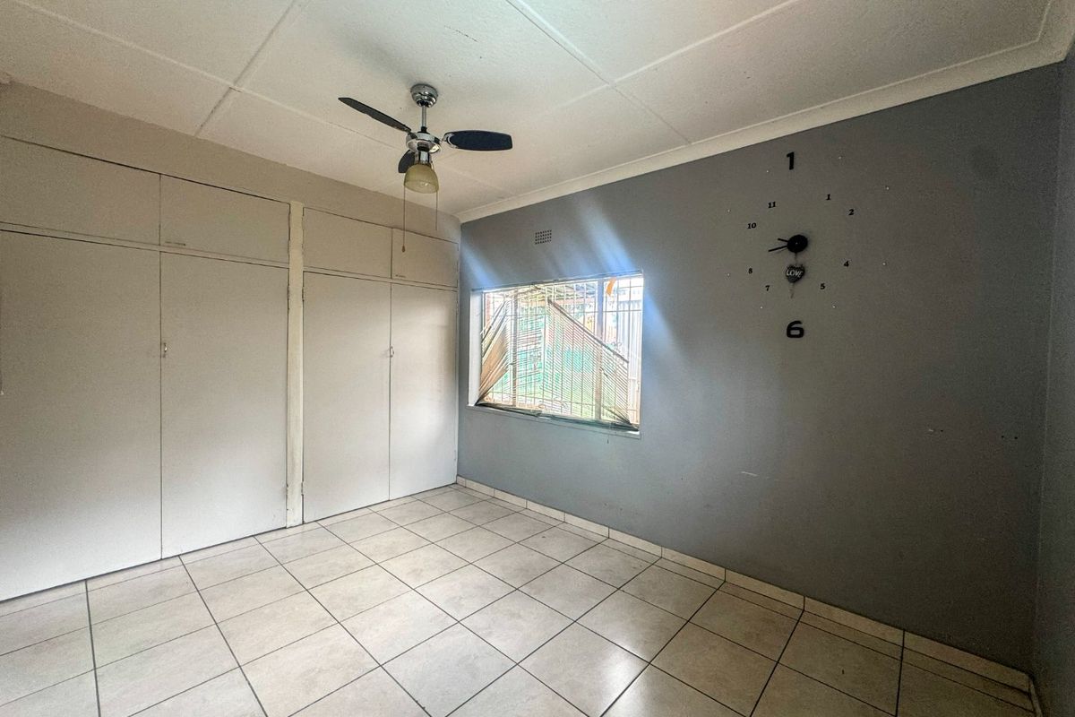 A vacant bedroom featuring tiled flooring, built-in cupboards, a ceiling fan, and a window fitted with security bars.