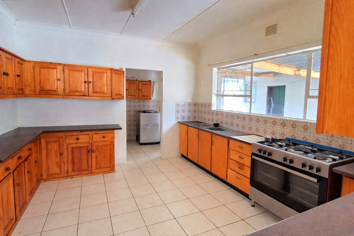 A functional kitchen featuring wooden cabinetry, a gas stove with oven, tiled flooring, and a tiled backsplash, with an adjacent scullery area.