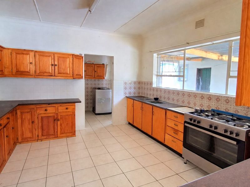 A functional kitchen featuring wooden cabinetry, a gas stove with oven, tiled flooring, and a tiled backsplash, with an adjacent scullery area.