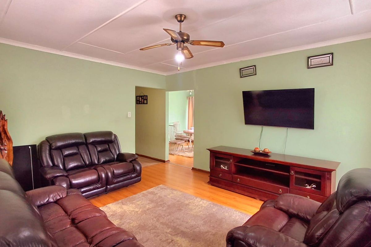 A spacious living room featuring laminate flooring, a ceiling fan, and neutral green walls, with an open-plan flow leading to the dining area.