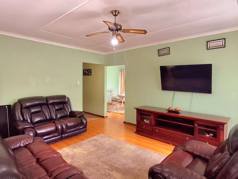 A spacious living room featuring laminate flooring, a ceiling fan, and neutral green walls, with an open-plan flow leading to the dining area.