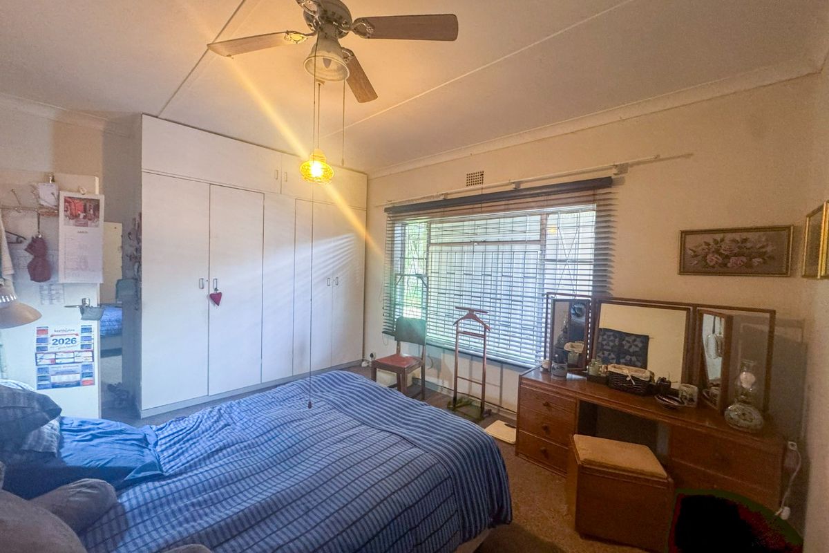 A functional bedroom featuring built-in cupboards, a ceiling fan, and large windows with venetian blinds, offering a comfortable and practical living space.