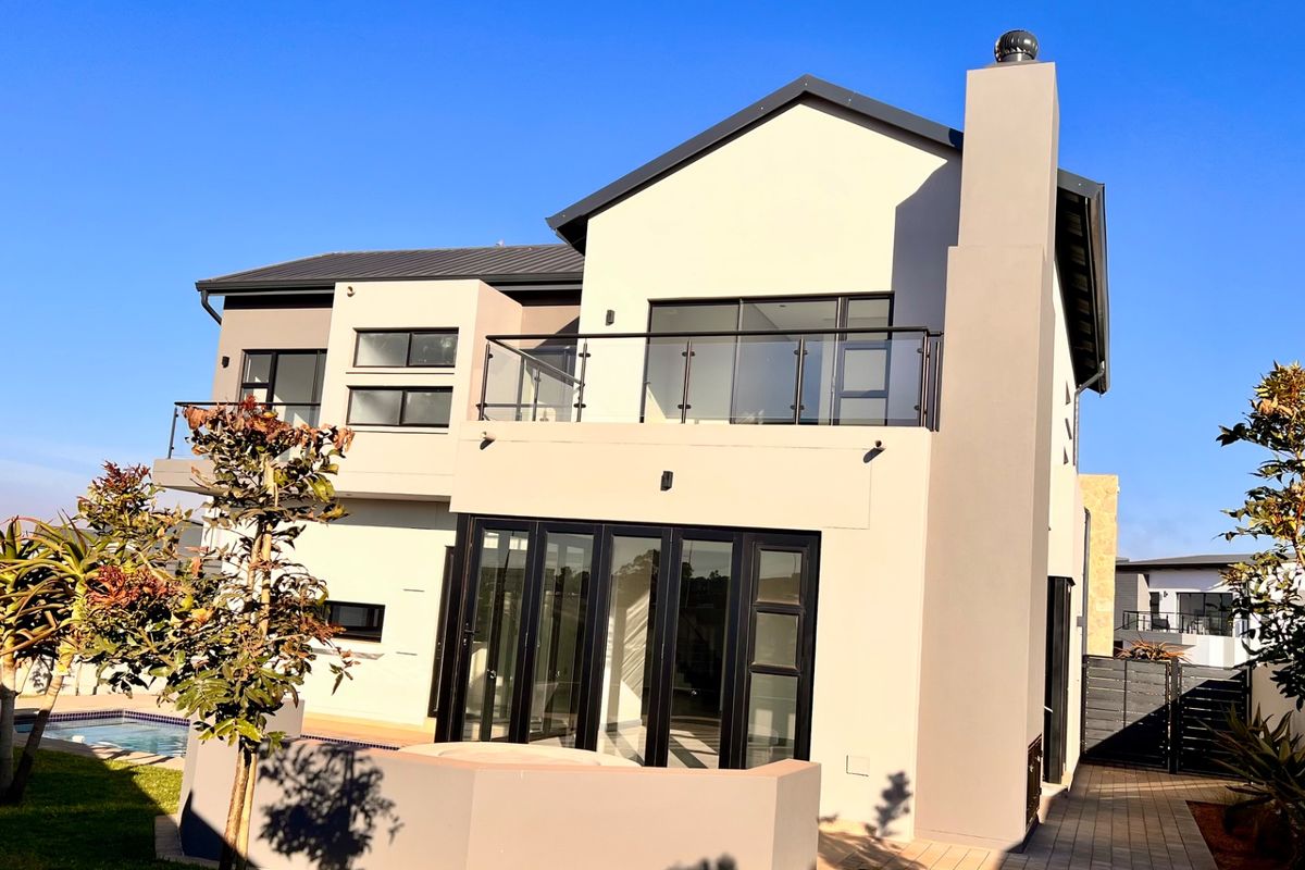 Rear view of a modern double-story house featuring a balcony with glass balustrades, bi-fold doors, a chimney, and a swimming pool in the garden.