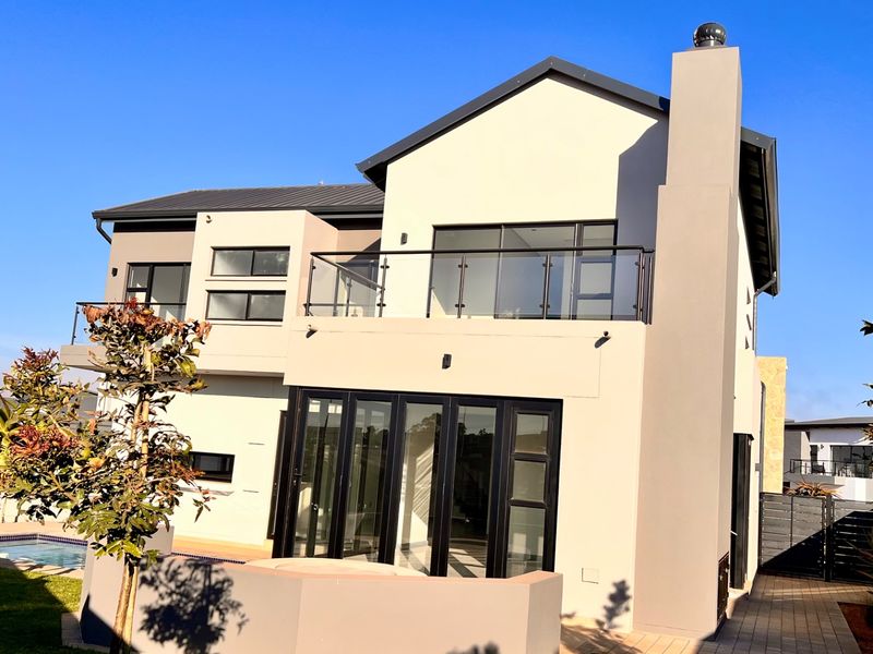 Rear view of a modern double-story house featuring a balcony with glass balustrades, bi-fold doors, a chimney, and a swimming pool in the garden.
