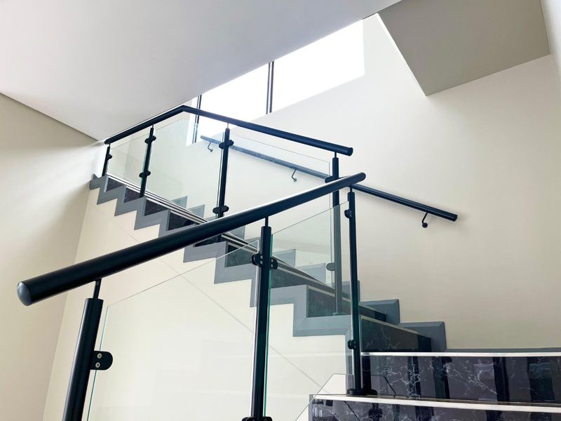 Modern staircase featuring a sleek glass balustrade, black metal handrails, and marble-look steps, illuminated by a bright overhead skylight.