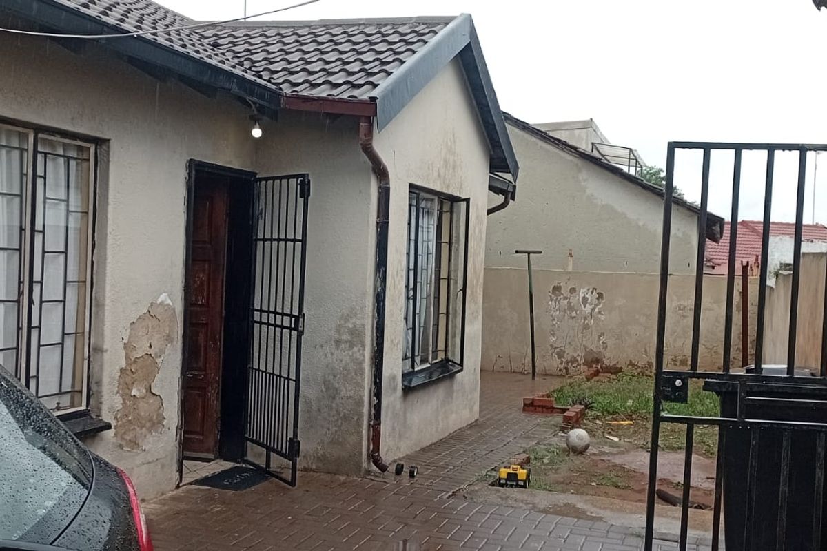 Rear exterior view of the house featuring a paved area, security gate, and tiled roof, showing signs of wear on the exterior wall finishes.