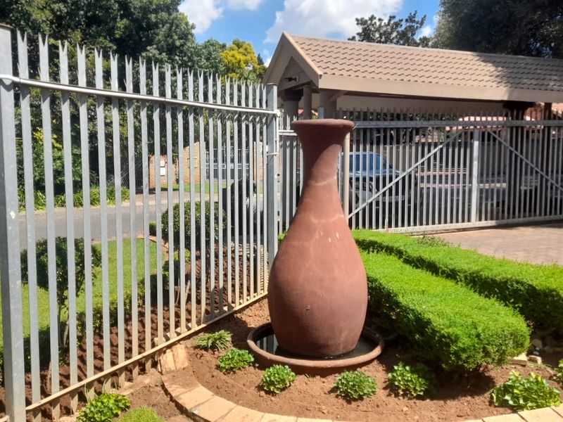 Front garden area featuring a decorative water feature, steel palisade fencing, and a paved driveway leading to a covered carport.