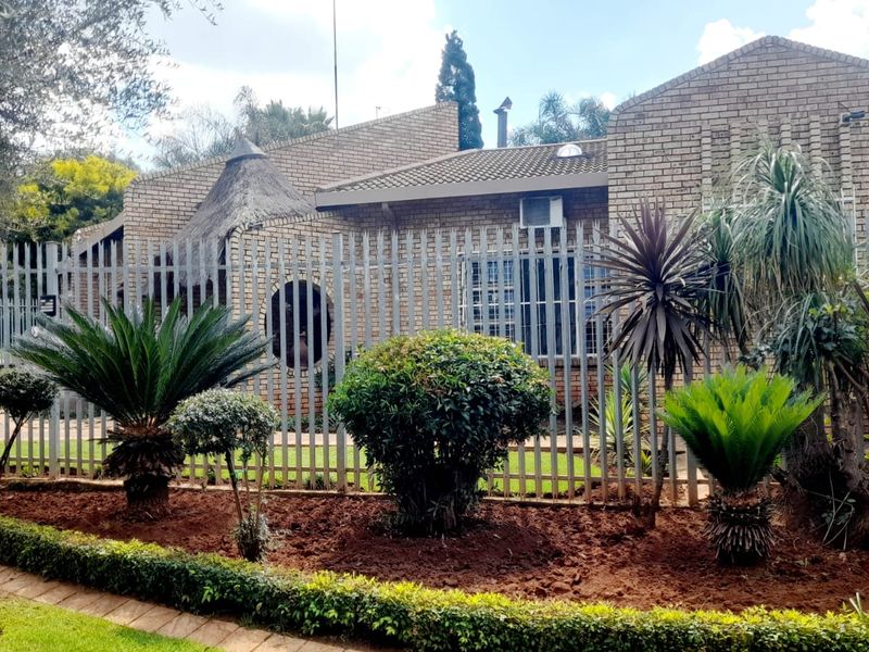 Front elevation of a face brick house featuring a unique thatched roof section, secure steel fencing, and well-maintained landscaped garden beds.