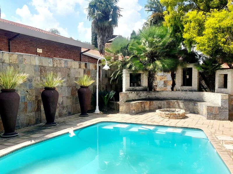A well-maintained outdoor pool area featuring a stone-clad boundary wall, built-in stone seating, a central fire pit, and paved decking.