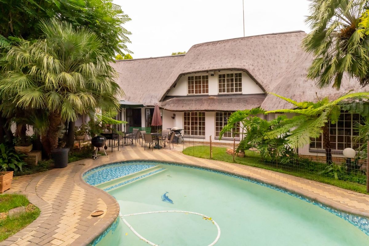 Rear view of a thatched-roof house featuring a paved patio area and a swimming pool set within a landscaped garden.