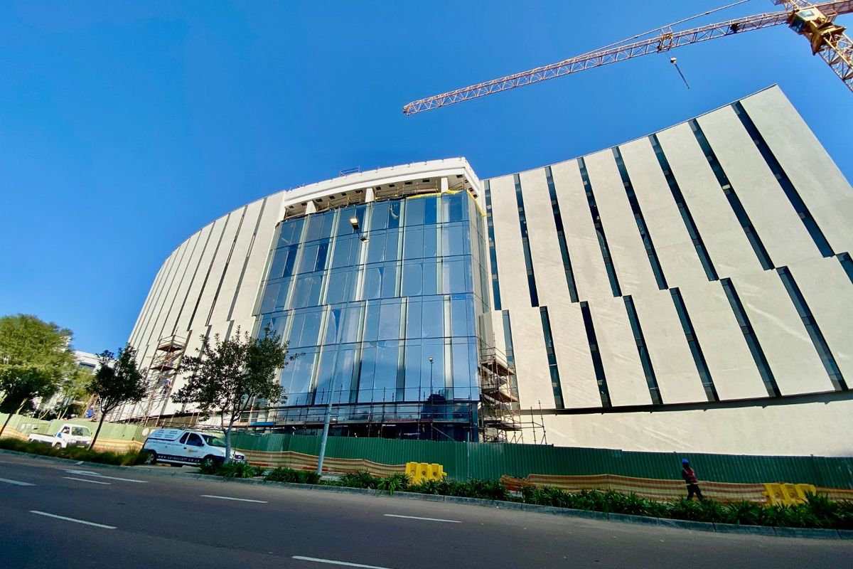 A large commercial building under construction featuring a prominent glass curtain wall facade and concrete exterior elements.