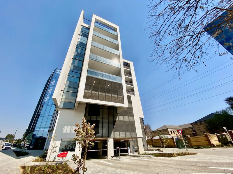 Modern multi-story commercial building featuring a glass facade, integrated balconies, and a secure ground-level parking entrance with paved access.