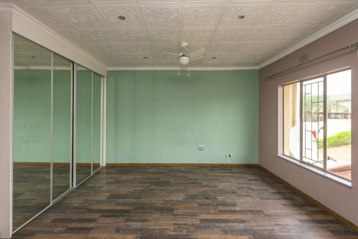 A spacious, empty bedroom featuring laminate flooring, a large mirrored sliding wardrobe, decorative ceiling tiles, and a ceiling fan.