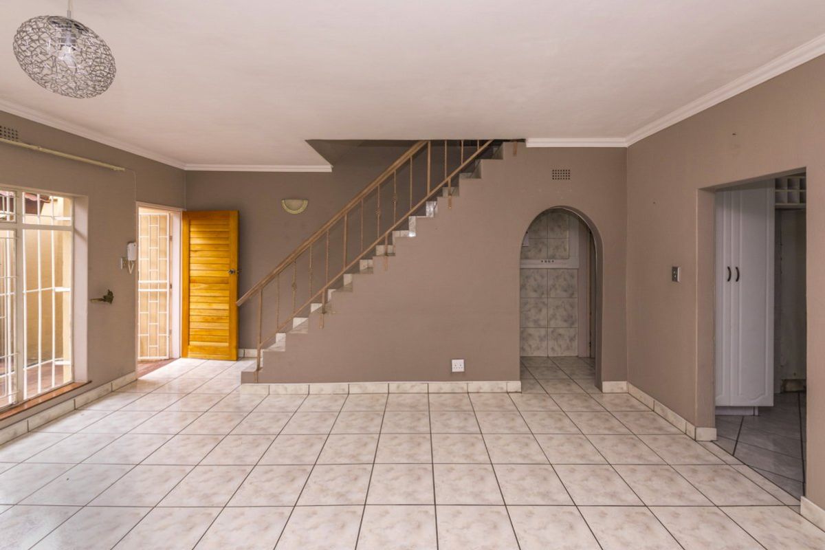 An open-plan living area featuring tiled flooring, a central staircase with metal railing, and an arched entryway leading to adjacent spaces.