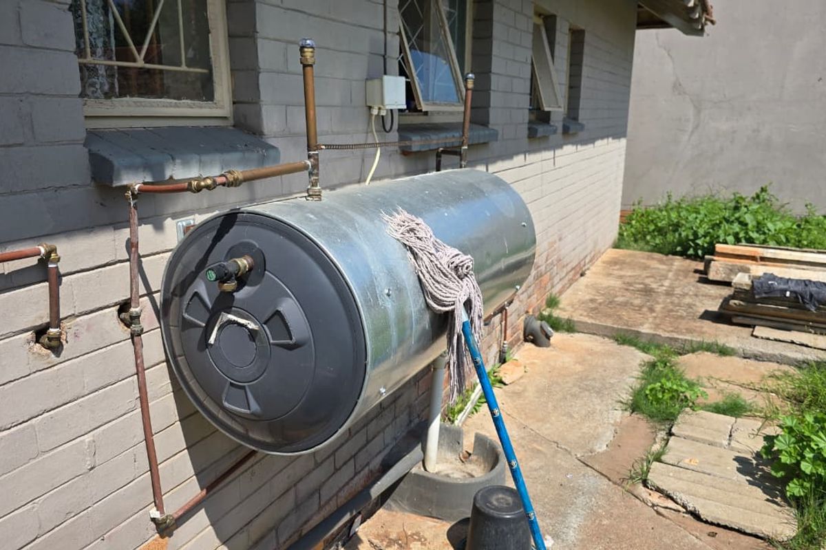 Exterior view of a wall-mounted electric water heater unit with exposed copper piping and brick facade.