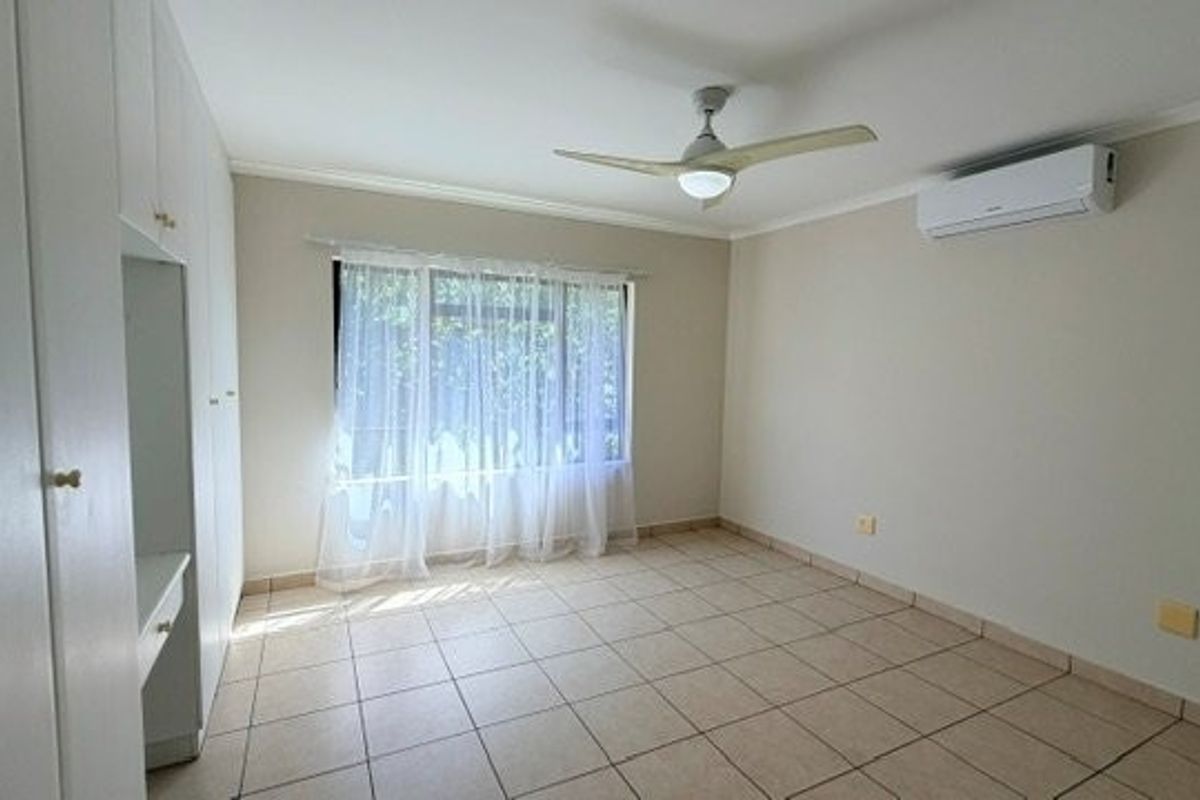 A bright, empty bedroom featuring tiled flooring, built-in cupboards, a ceiling fan, and a wall-mounted air conditioning unit.