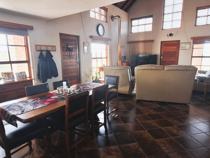 Spacious open-plan living and dining area featuring vaulted ceilings, tiled floors, and a wood-burning fireplace with abundant natural light from large timber-framed windows.