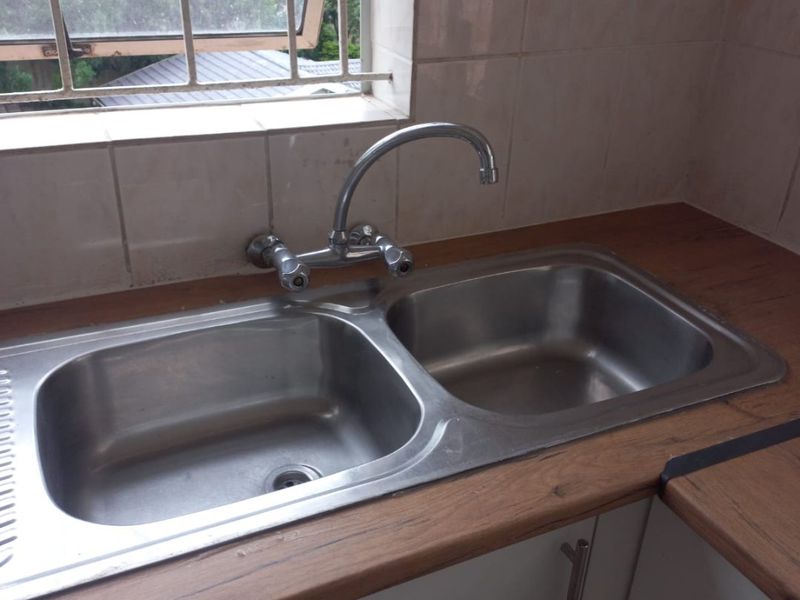 A functional kitchen sink area featuring a double bowl stainless steel basin, laminate countertop, and tiled splashback with security bars on the window.