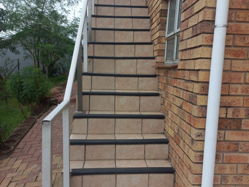 Exterior view of a tiled staircase with a metal handrail leading to an upper level, featuring facebrick construction and a paved walkway.