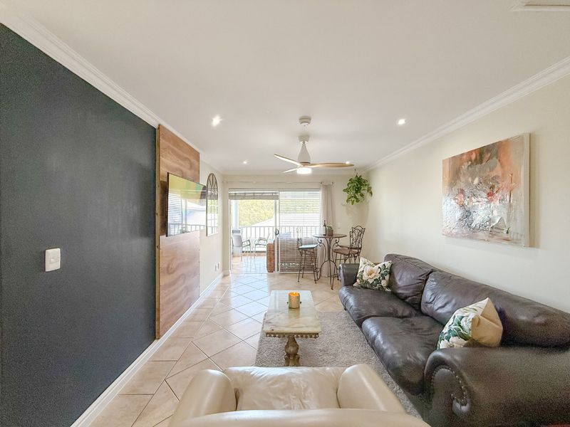 A well-lit living room featuring tiled flooring, a ceiling fan, and an accent wall, with direct access to an outdoor balcony through sliding glass doors.