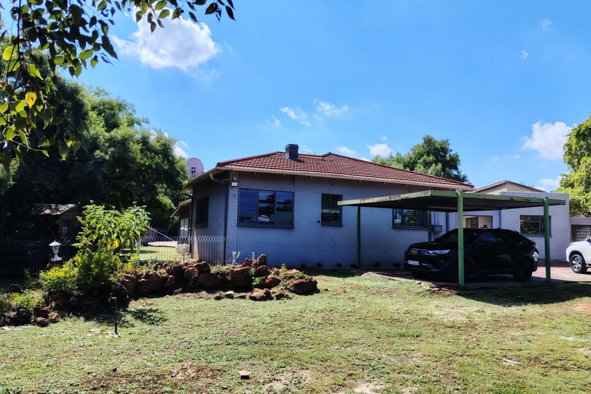 Rear view of a single-story brick house featuring a tiled roof, chimney, and an attached carport structure set within a garden area.
