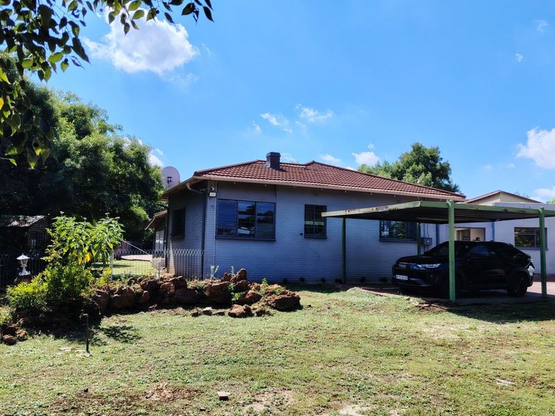 Rear view of a single-story brick house featuring a tiled roof, chimney, and an attached carport structure set within a garden area.
