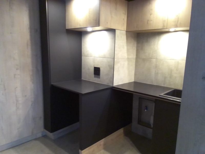 A modern, compact scullery or utility area featuring sleek dark cabinetry, stone countertops, integrated sink, and stylish tiled splashback with under-cabinet lighting.