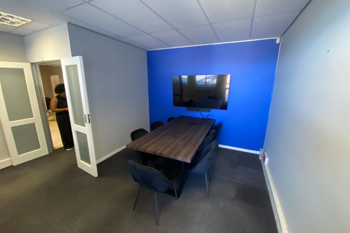 A functional meeting room featuring a dark wood-look table, wall-mounted display screen, suspended ceiling tiles, and a bold blue accent wall.