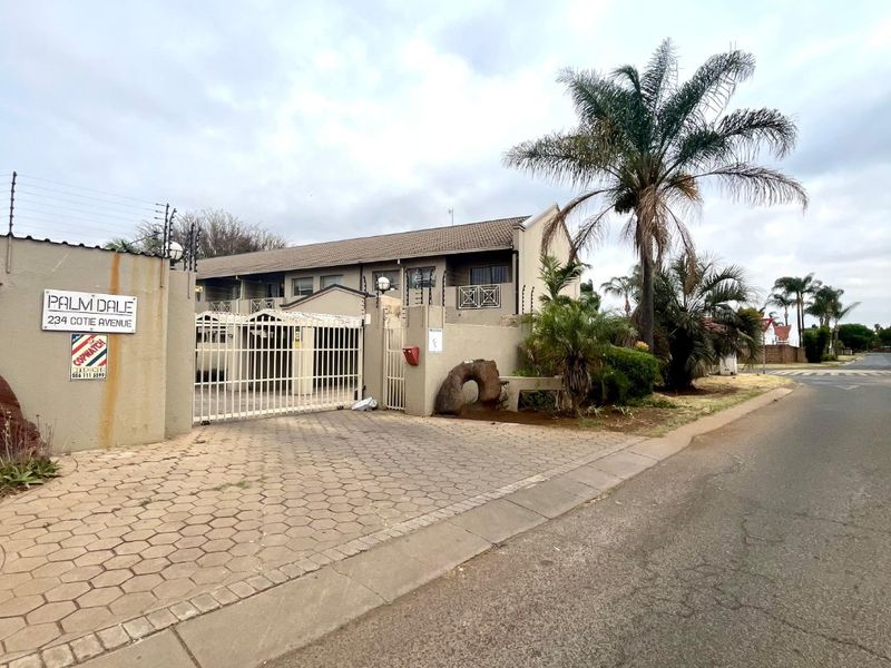 Front exterior view of Palm Dale townhouse complex featuring a paved driveway, security gate, and perimeter wall with electric fencing.