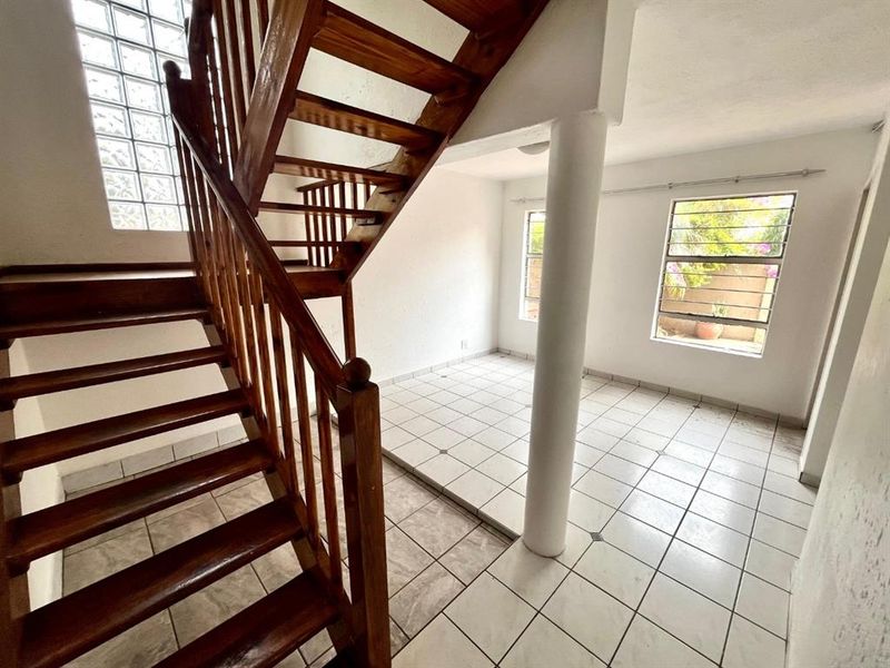 An open-plan living area featuring a prominent wooden staircase, ceramic tiled flooring, a structural column, and natural light from glass block and casement windows.