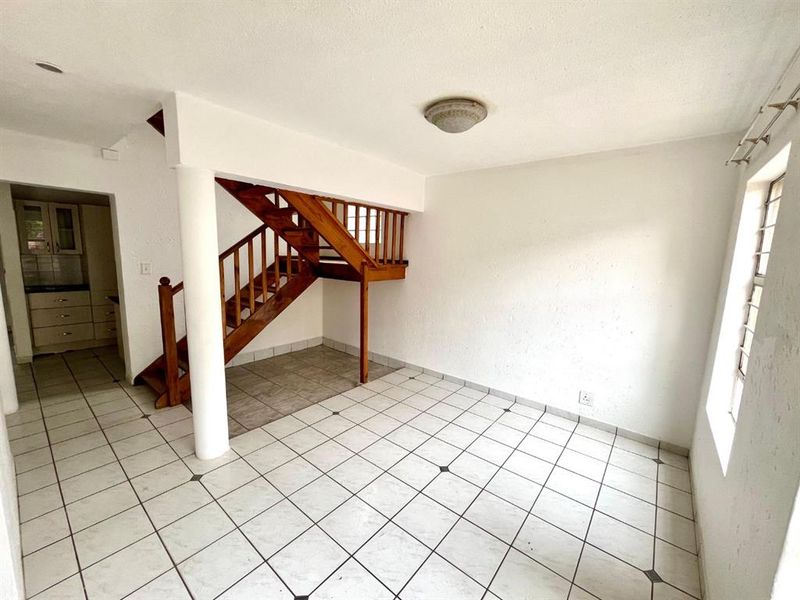 An empty living area featuring ceramic floor tiles, a wooden staircase, and a structural column, leading through to a kitchen area.